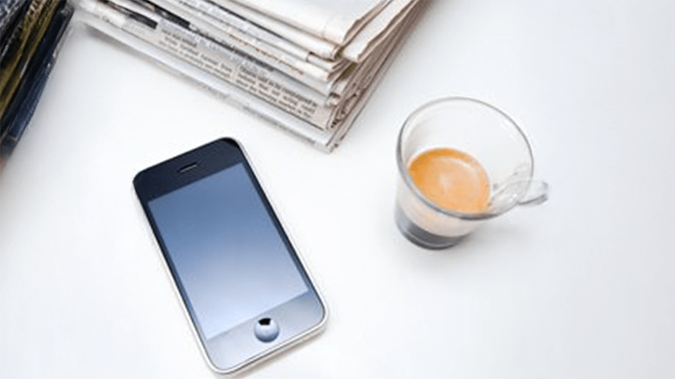 Iphone laying on table near a stack of newspapers and a glass of coffee.