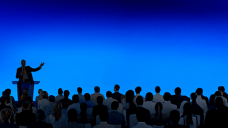 A man stands at a lectern on a stage in front of a crowd and speaks into a mic.