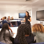 A women stands and teaches a classroom of students gathered around