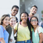 A group of eight multi-ethnic teenagers, 17 and 18 years old, carrying book bags, standing together outside a school building. They are high school seniors or university freshmen.