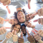 A group of people in matching shirts look down at a camera and give a thumbs up in a circle