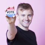 A blonde man holds a sign to the camera that reads 'VOTE' in red, white, and blue