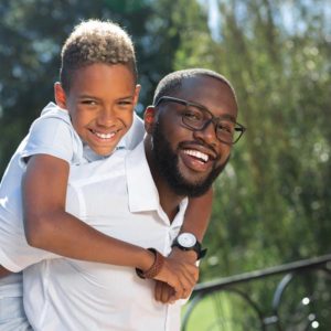 A black man smiles as a young boy behind him embraces him around his neck